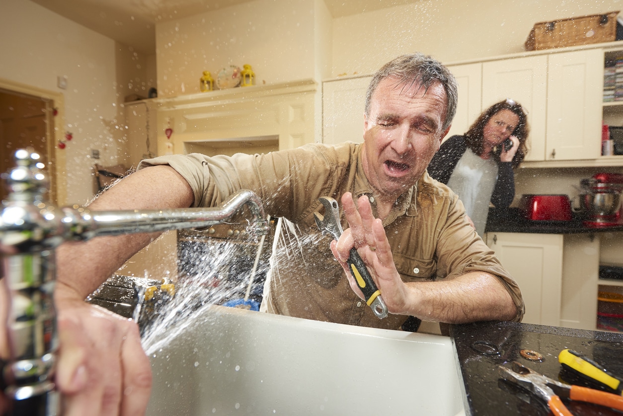 Image of a water from a sink spraying a man, while a concerned woman is on the phone in the background.